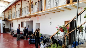 Amplio espacio interior con piso rojo, vigas de madera y barandales de metal. Se ven varias personas, algunas con equipaje, y plantas en macetas a lo largo de las paredes.