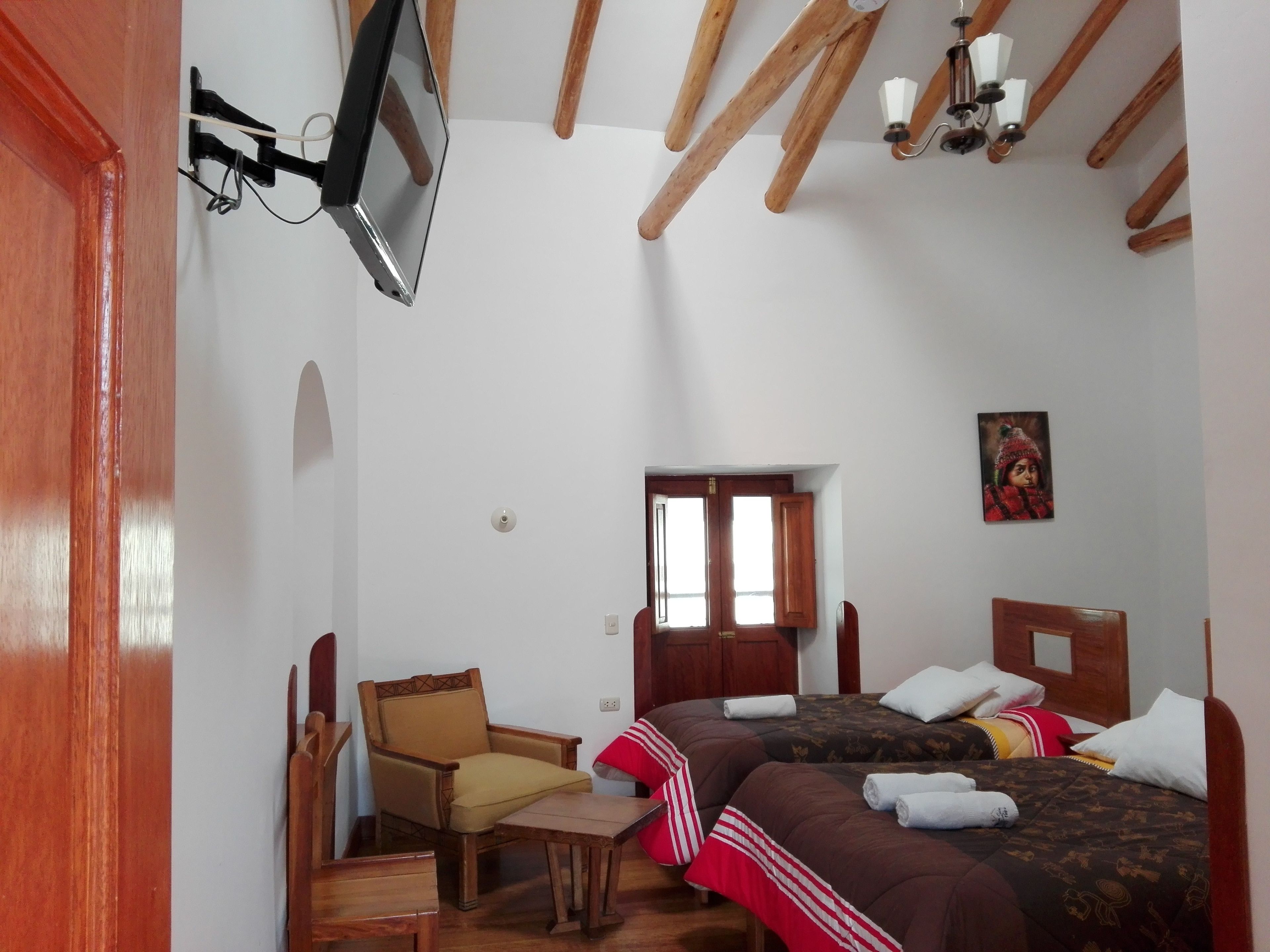 A compact room with two beds, a wooden chair, a small table, a TV mounted on the wall, and a ceiling light fixture.