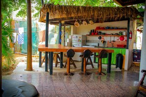 A rustic outdoor kitchen with a thatched roof, a dining table, and chairs. - Pura Vida Hostel (Tamarindo)