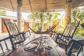 A wooden table with a monkey sculpture, surrounded by two wooden chairs, under a thatched roof with a hammock between them. - Pura Vida Hostel (Tamarindo)