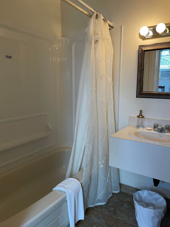A bathroom with a bathtub, a shower curtain, a vanity with a mirror, and a trash can. - Marshlands Inn (Sackville)