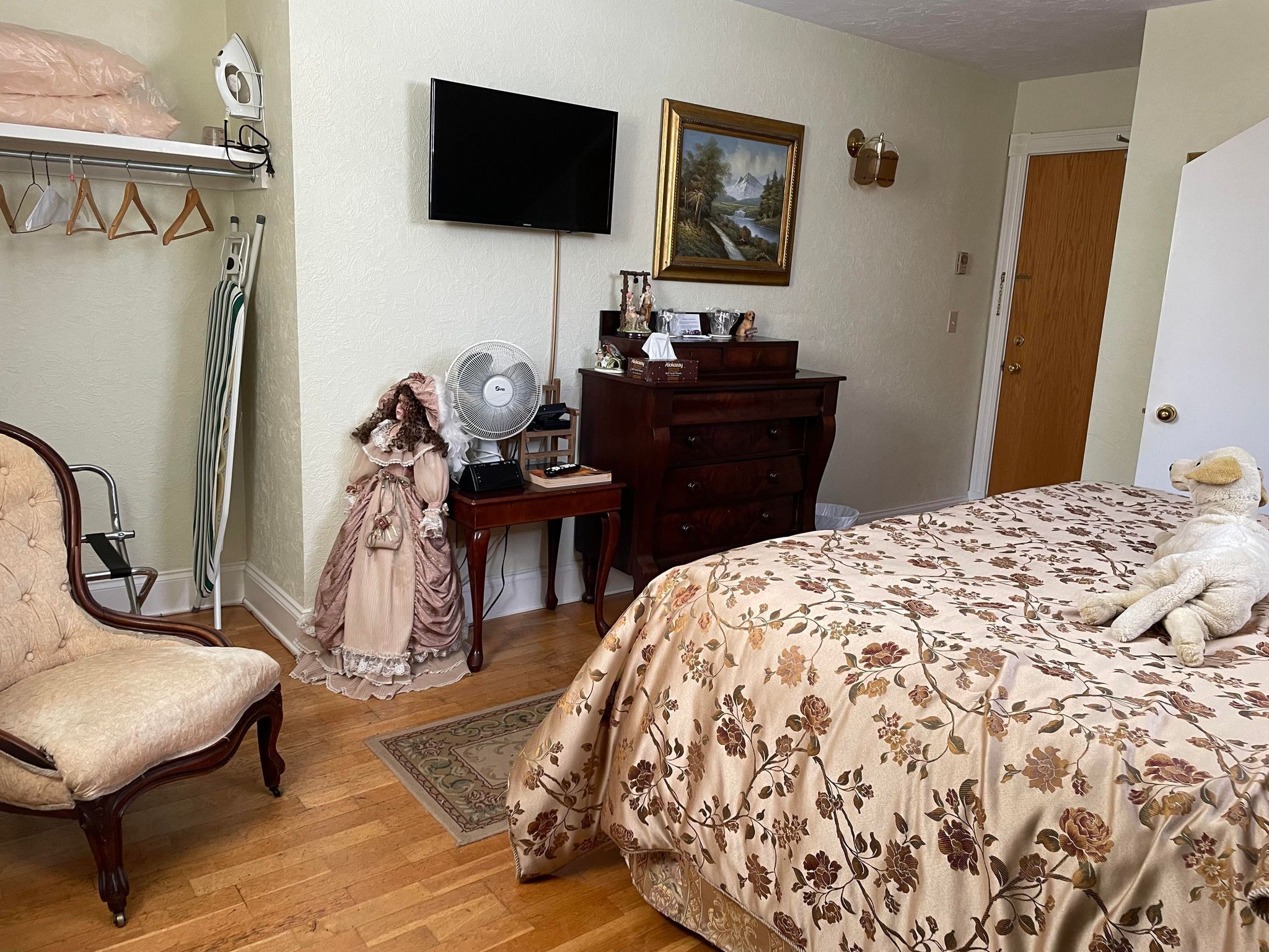 A bedroom with a wooden bed, a dresser, a television, a fan, a chair, and a doll.