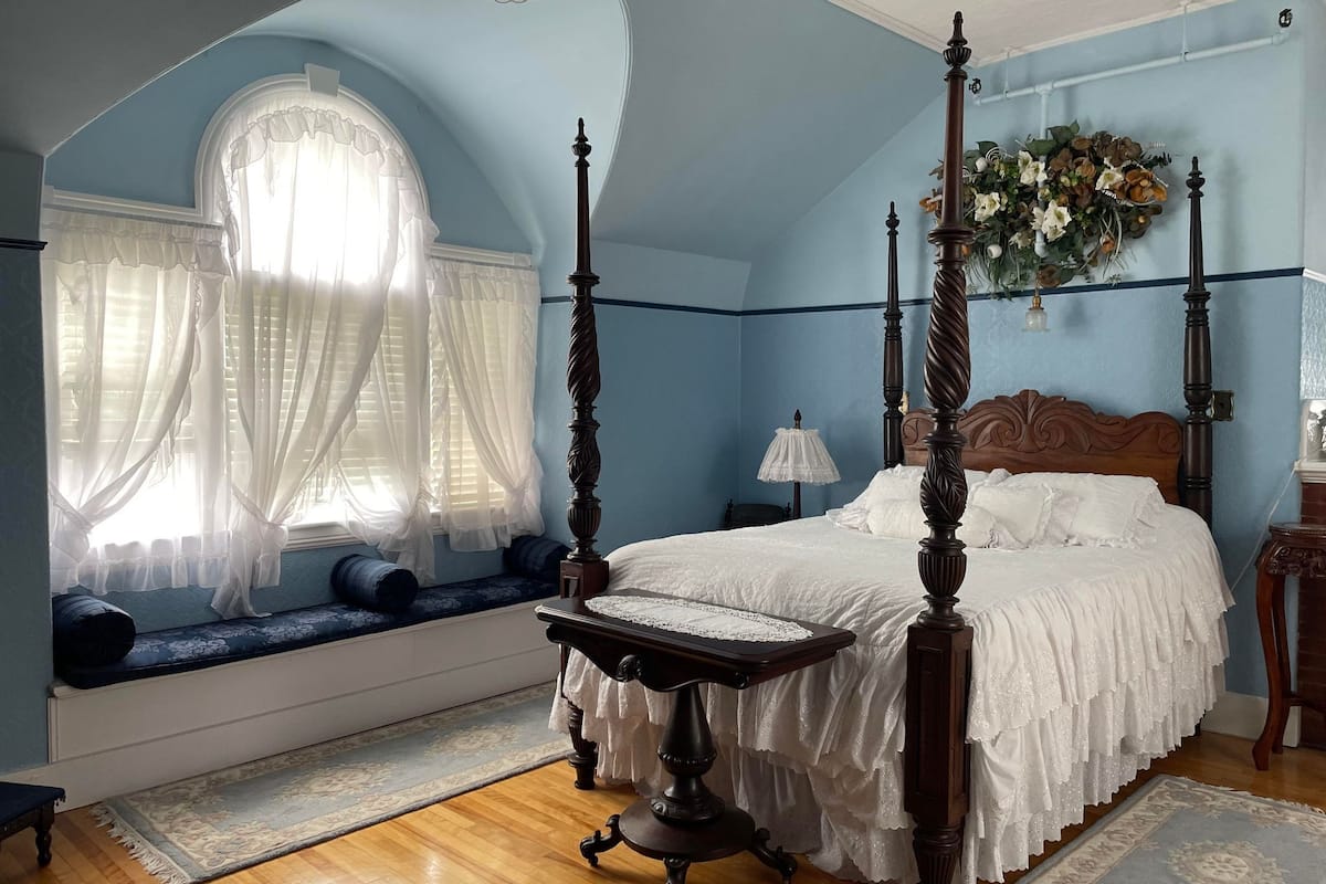 Un dormitorio con cama con dosel, una ventana con cortinas traslúcidas, un banco con cojines y un adorno floral en la pared.