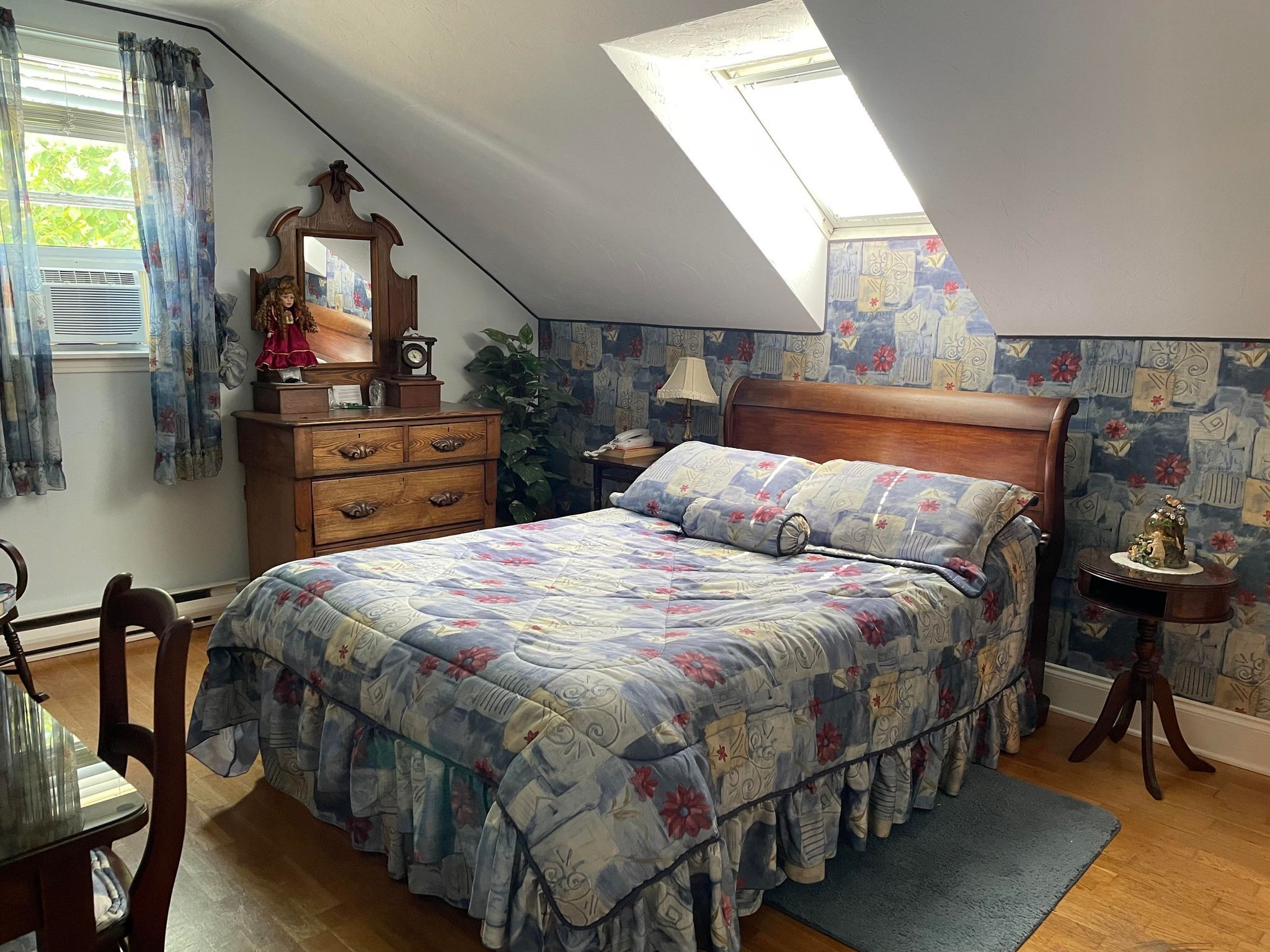 Una camera da letto con soffitto a falde, un letto con copriletto floreale, un comò in legno, uno specchio e un comodino con una lampada.