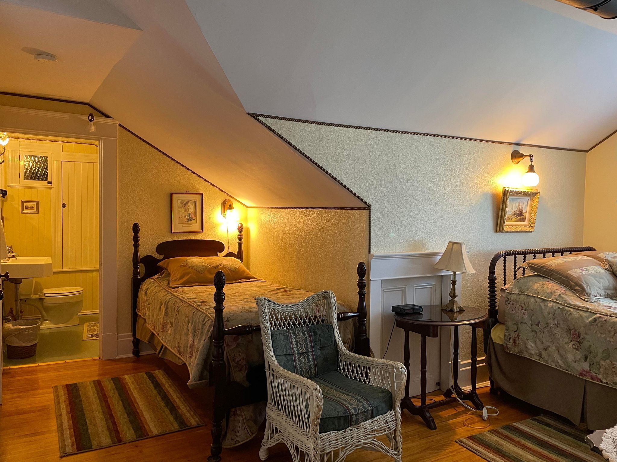A cozy bedroom with two beds, a wicker chair, a small table, and a bathroom visible through an open door.