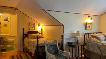 A cozy bedroom with two beds, a wicker chair, a small table, and a bathroom visible through an open door.