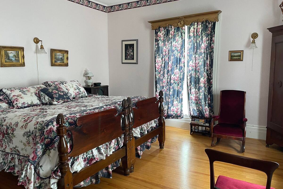 Un dormitorio con cama de madera, ropa de cama floral, una silla roja y una mesita de noche con lámpara.