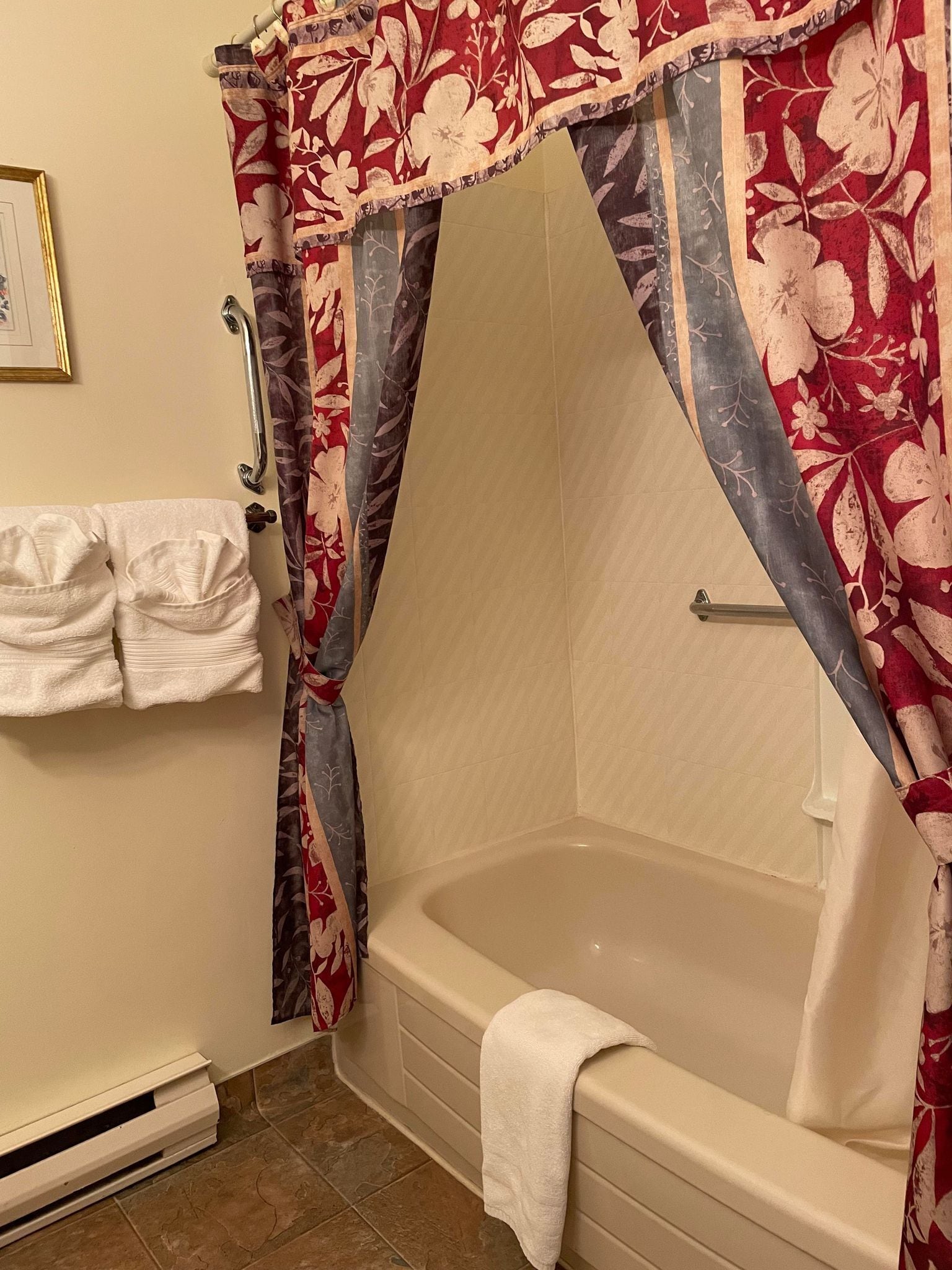 A bathroom with a bathtub, a shower curtain with a floral pattern, a towel rack with towels, and a handrail.