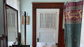 A room with a wooden floor, a white door with lace curtains, a mirror, a wooden chair, a small table with a green vase, and a patterned curtain. - Marshlands Inn (Sackville)