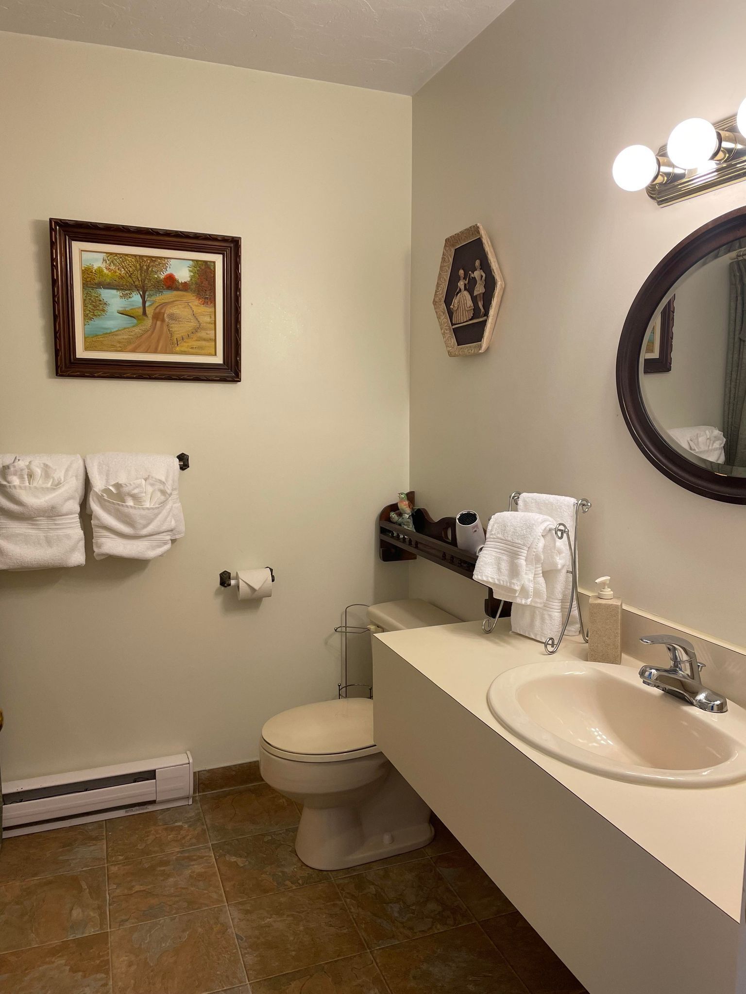 A bathroom with a toilet, sink, and towel rack. There is a framed picture on the wall and a mirror with lights.