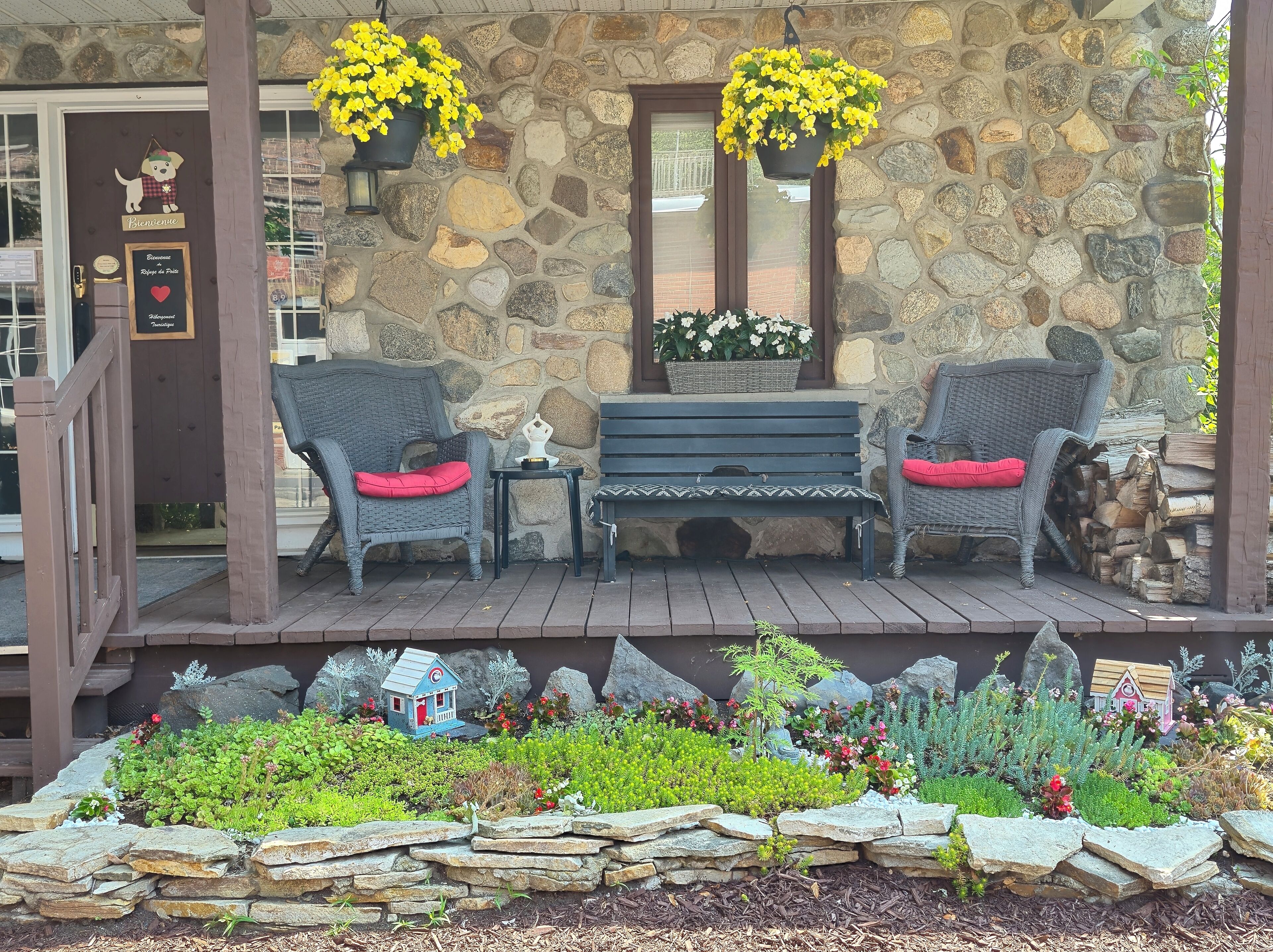 Un porche en pierre avec des fauteuils en osier et un banc, un petit jardin agrémenté d’une petite maison décorative et de jardinières suspendues.