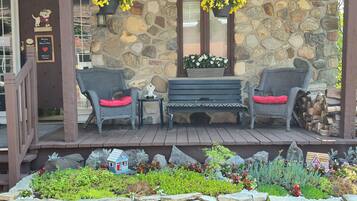 Un porche en pierre avec des fauteuils en osier et un banc, un petit jardin agrémenté d’une petite maison décorative et de jardinières suspendues.