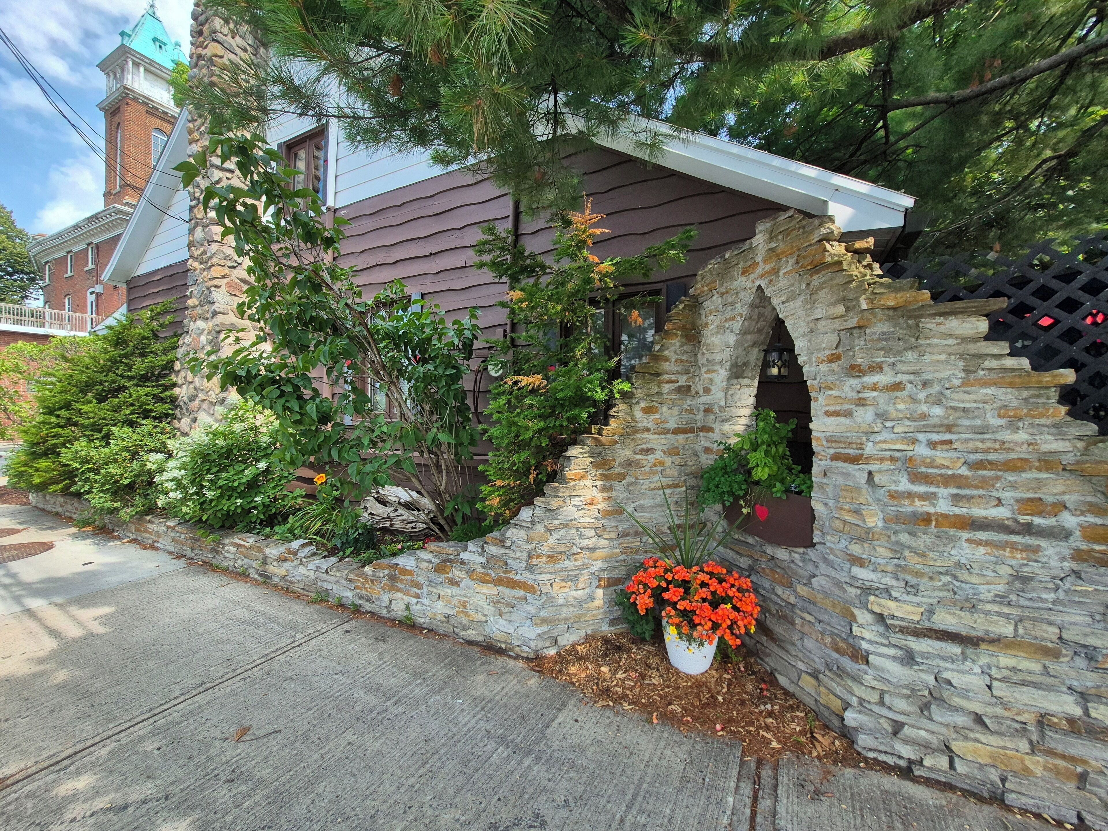 Une arche en pierre surmontée d’un pot de fleurs, un bâtiment en briques avec un toit végétalisé et un arbre au premier plan.
