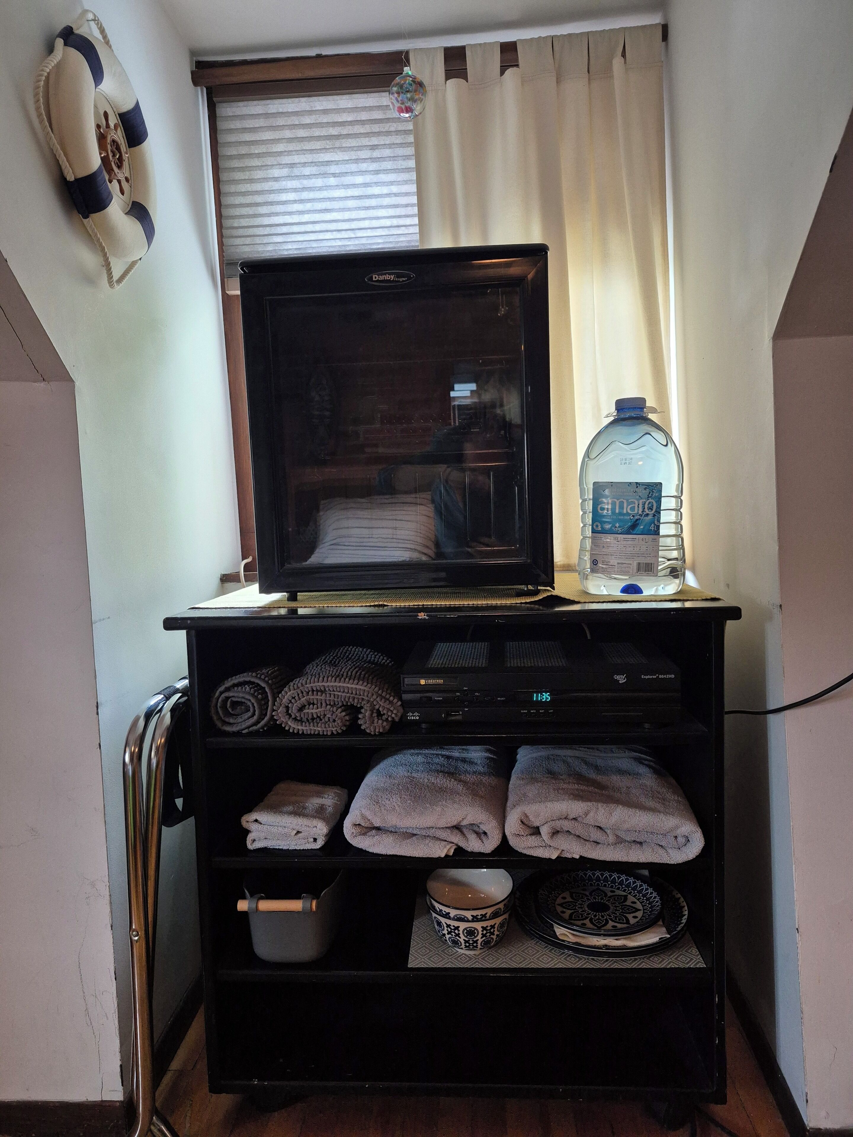 Une petite pièce avec un téléviseur posé sur un meuble, une bouteille d’eau, des serviettes, un bol et une assiette décorative.