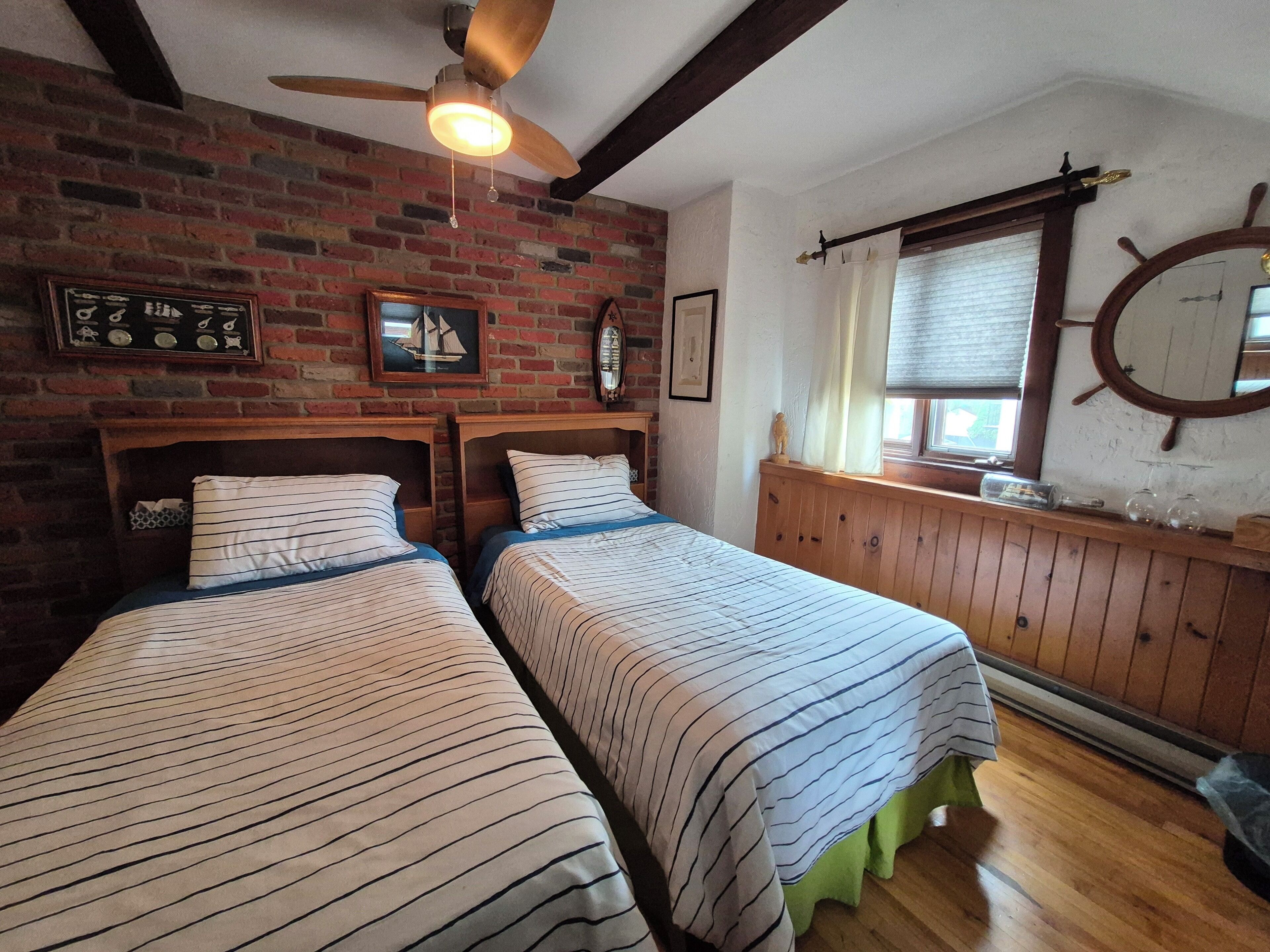 Une chambre avec deux lits, un ventilateur de plafond en bois, un mur en briques, une fenêtre avec des stores et un gouvernail de bateau accroché au mur.