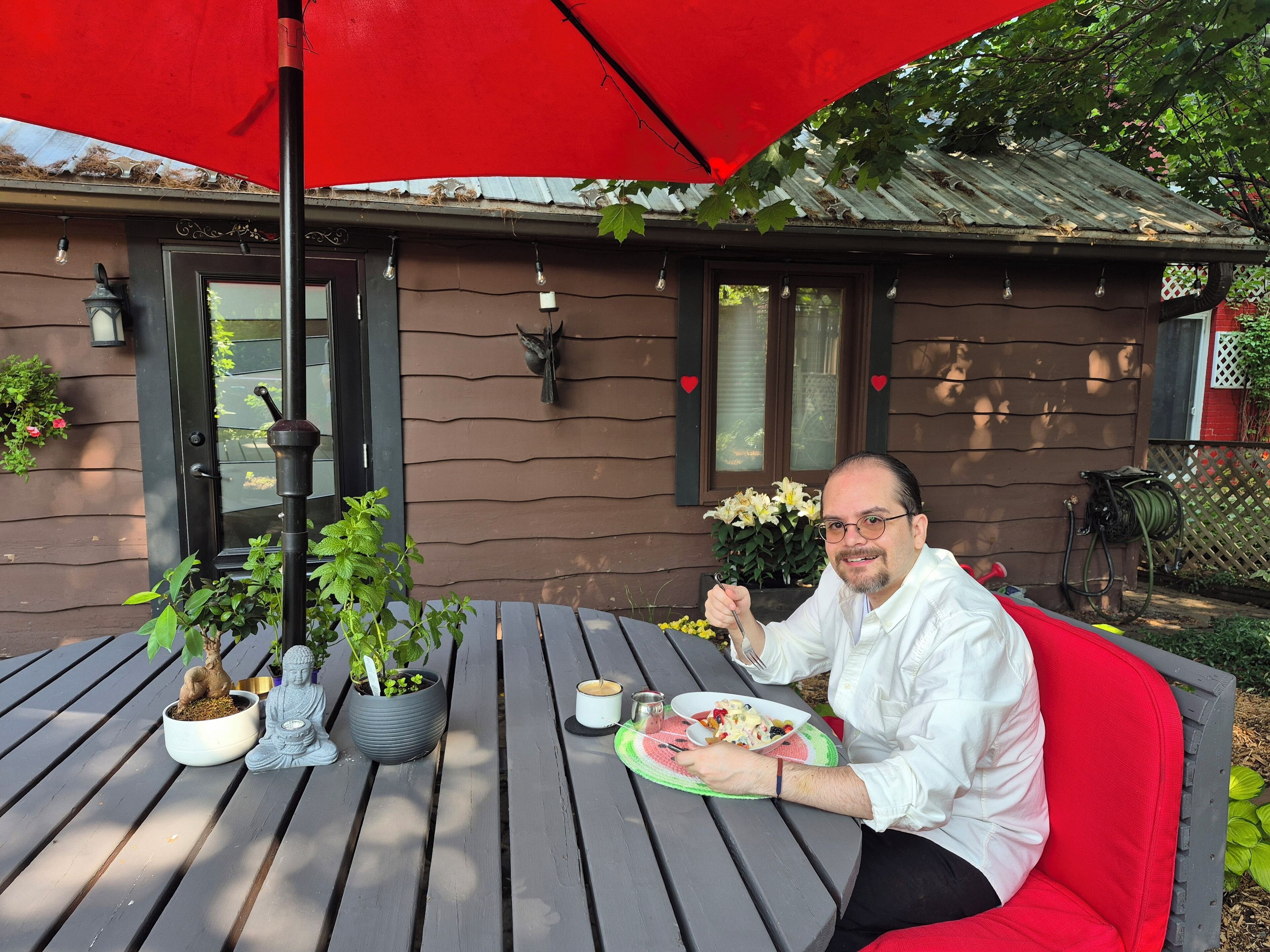 A man dining at an outdoor table with a red umbrella, surrounded by potted plants and a rustic building in the background.