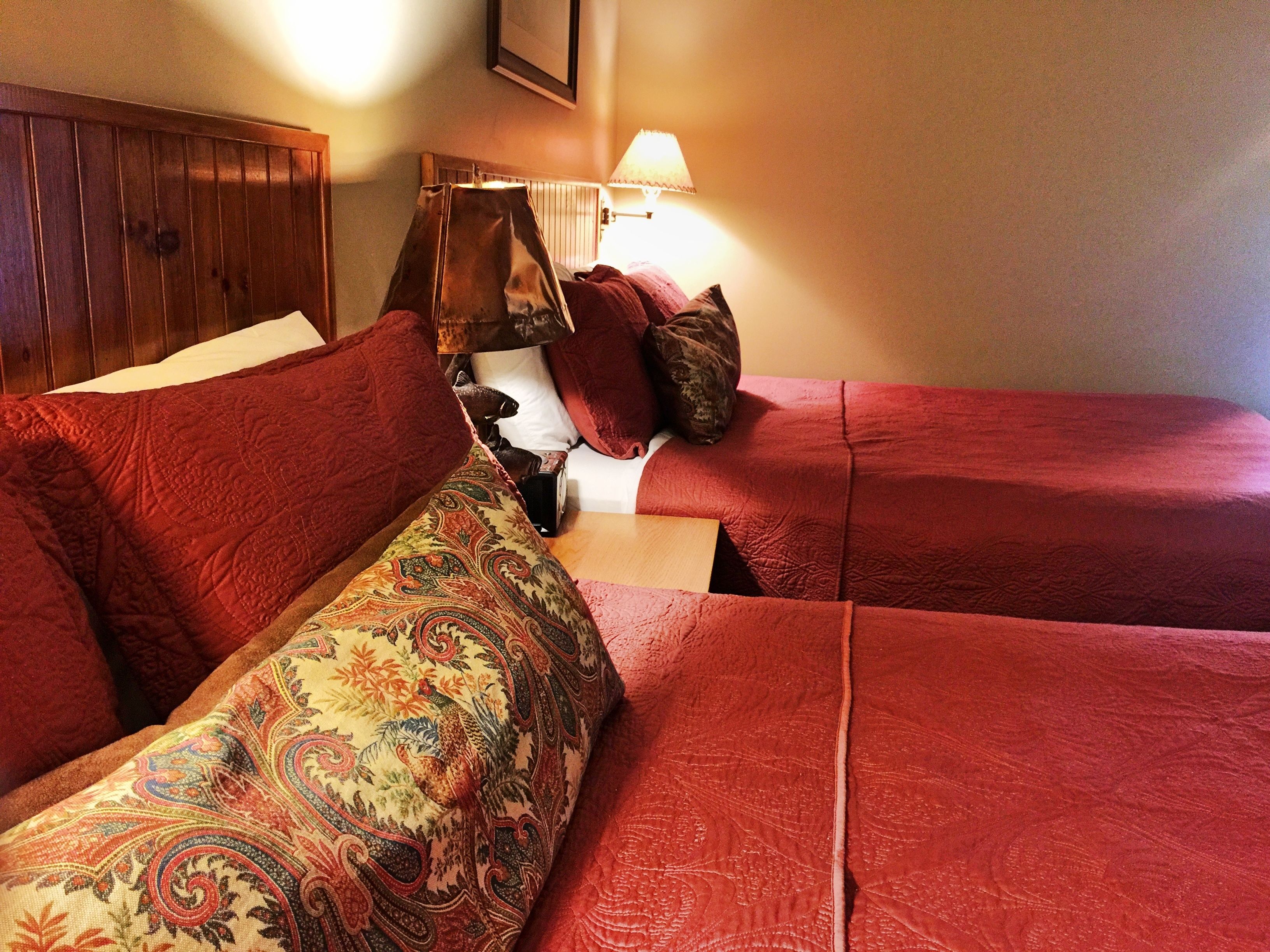 Une chambre d’hôtel avec deux lits, chacun agrémenté de draps rouges et d’un coussin décoratif à motifs. On y trouve une tête de lit en bois, une lampe et un tableau encadré au mur.