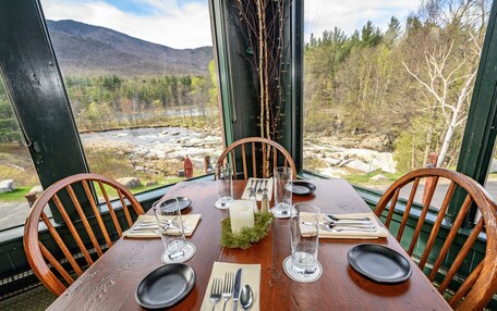 A wooden dining table set for four with a candle centerpiece, surrounded by chairs, overlooking a mountainous landscape with a river.