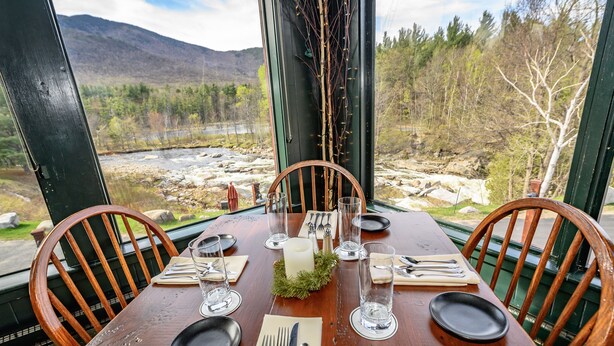 Une table à manger en bois, dressée pour quatre personnes, agrémentée d’un chandelier en guise de centre de table, entourée de chaises et offrant une vue sur un paysage montagneux traversé par une rivière.
