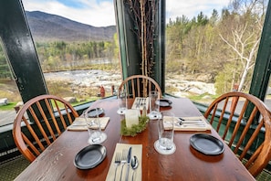 A wooden dining table set for four with a candle centerpiece, surrounded by chairs, overlooking a mountainous landscape with a river. - The Hungry Trout (Wilmington)
