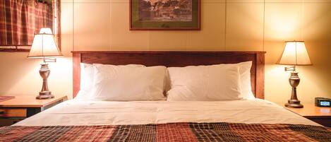 A neatly made bed with white linens, flanked by two bedside lamps, in a room with a large framed landscape painting above the headboard.