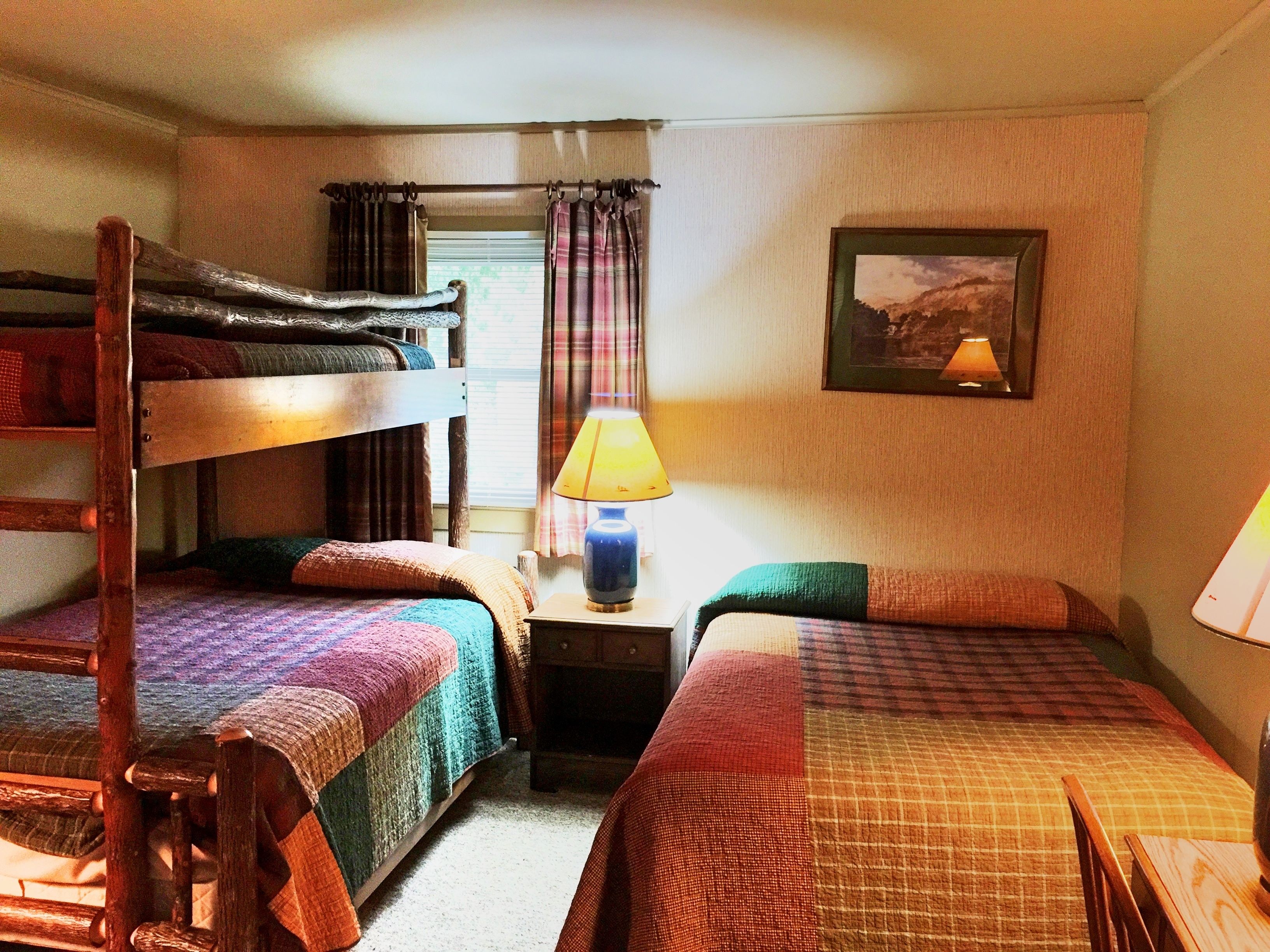 A room with bunk beds, a bedside table with a lamp, a framed picture on the wall, and a window with curtains.