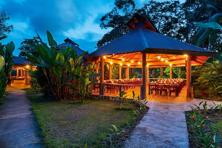 A covered outdoor dining area with tables and chairs, surrounded by tropical vegetation.