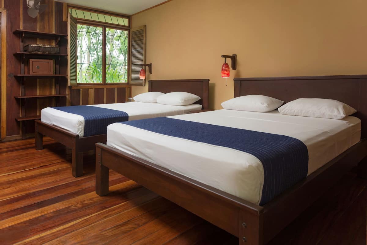 Two wooden beds with white and blue bedding, a wooden bench, a window with shutters, and a wall-mounted lamp.