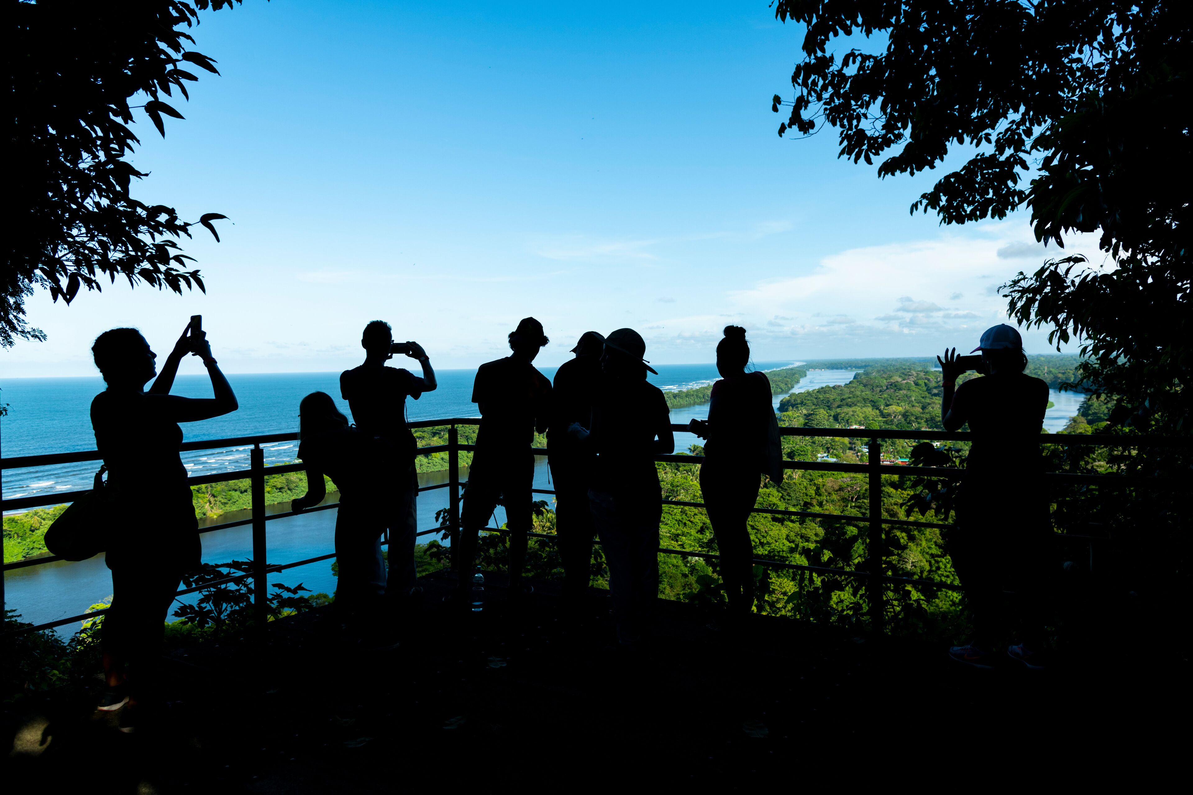 Silhouettes of people standing by a railing overlooking a body of water and greenery.