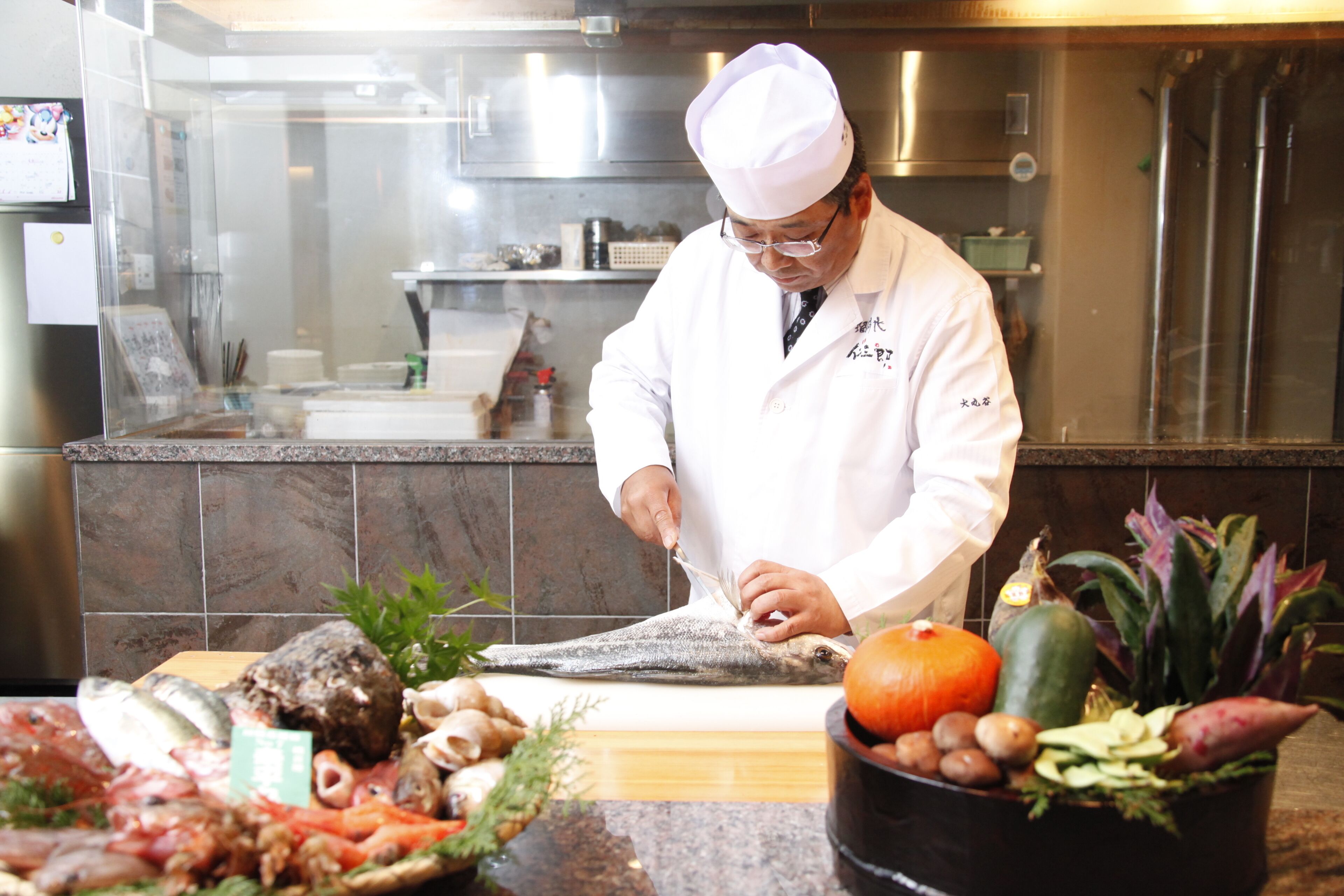 A chef in a white uniform preparing food in a kitchen with stainless steel appliances and a tiled backsplash.