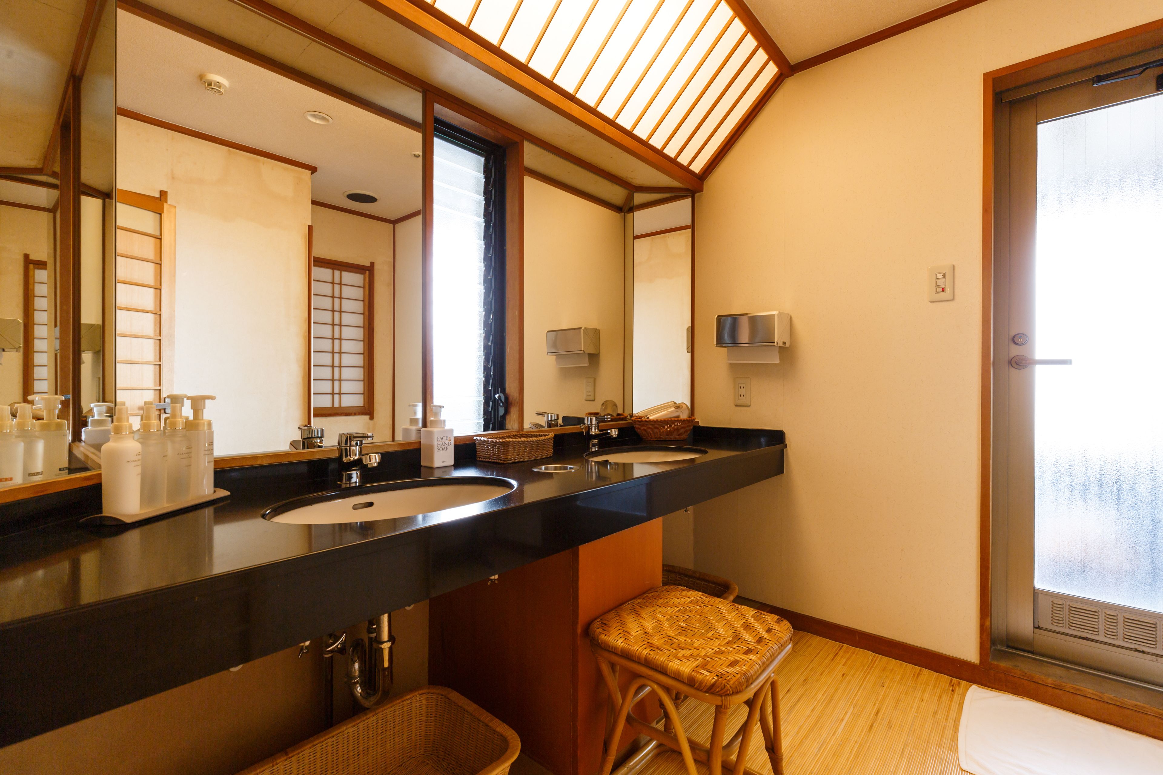 Une salle de bain avec un grand miroir, deux lavabos, un tabouret en osier et une porte coulissante en verre.