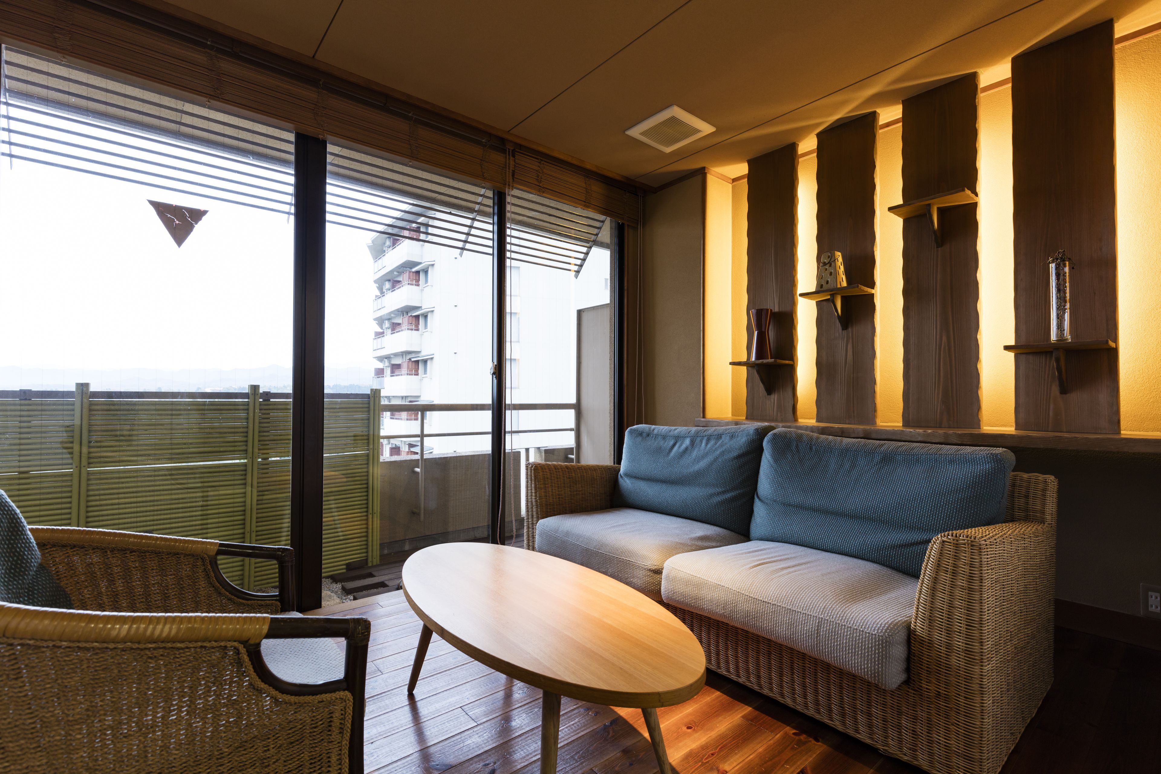 A balcony with a wicker sofa and a small table, wooden flooring, and a view of a building through the glass doors.