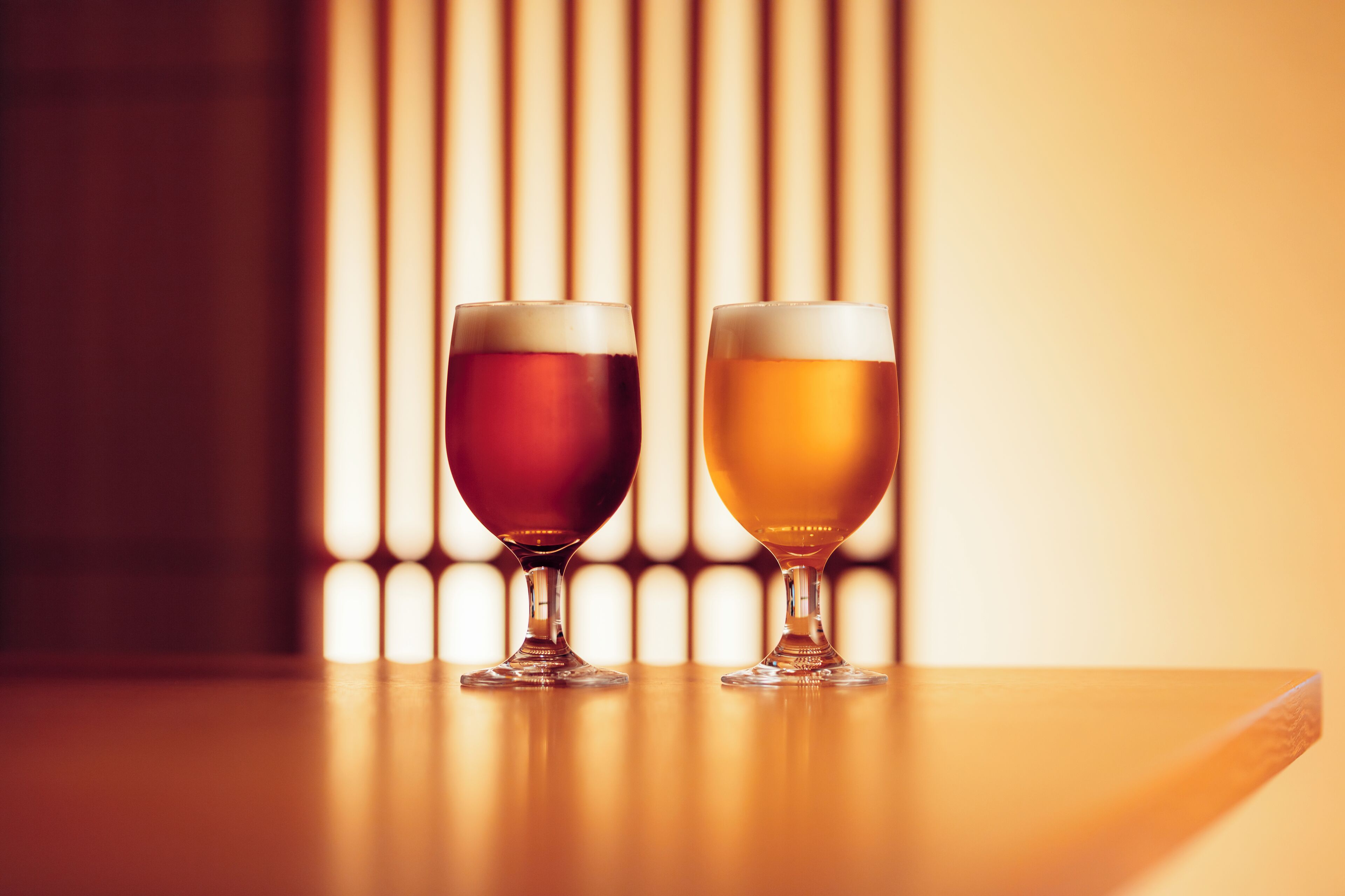 Two glasses of beer on a wooden table with vertical blinds in the background.