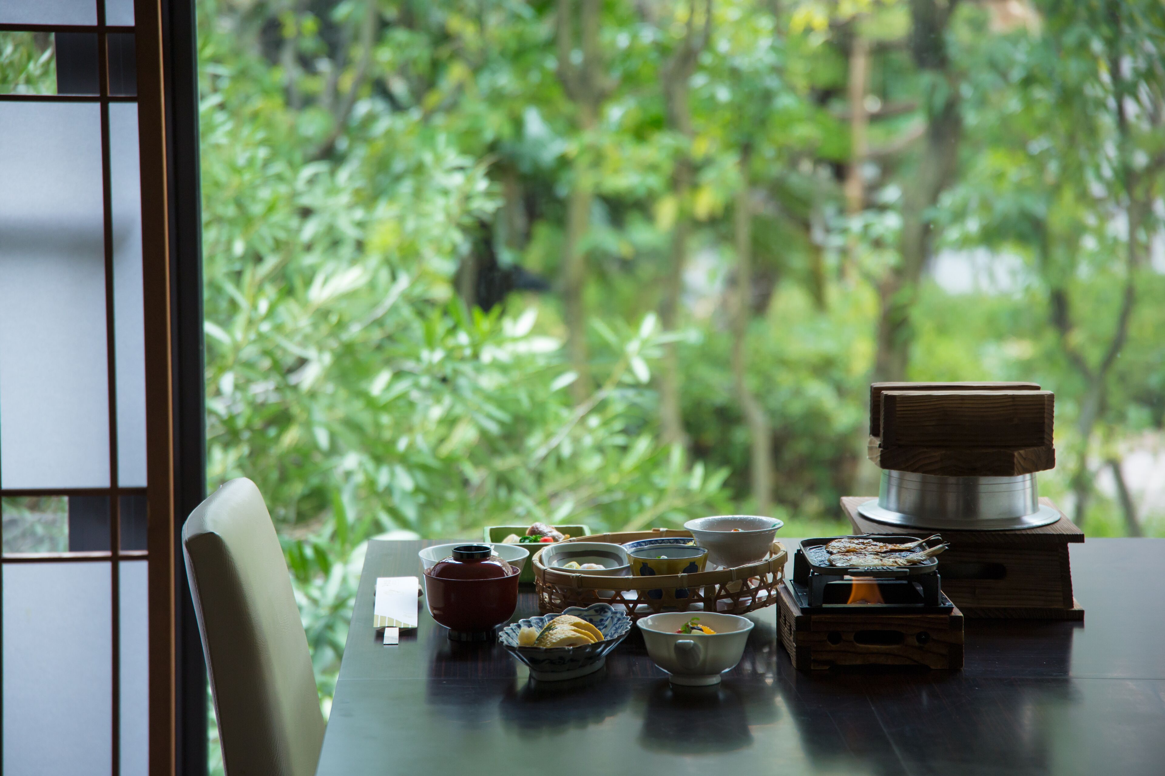 Una mesa con diferentes recipientes de té y café, una pequeña estufa y vistas de exuberante vegetación a través de una ventana.