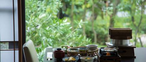Una mesa con diferentes recipientes de té y café, una pequeña estufa y vistas de exuberante vegetación a través de una ventana.
