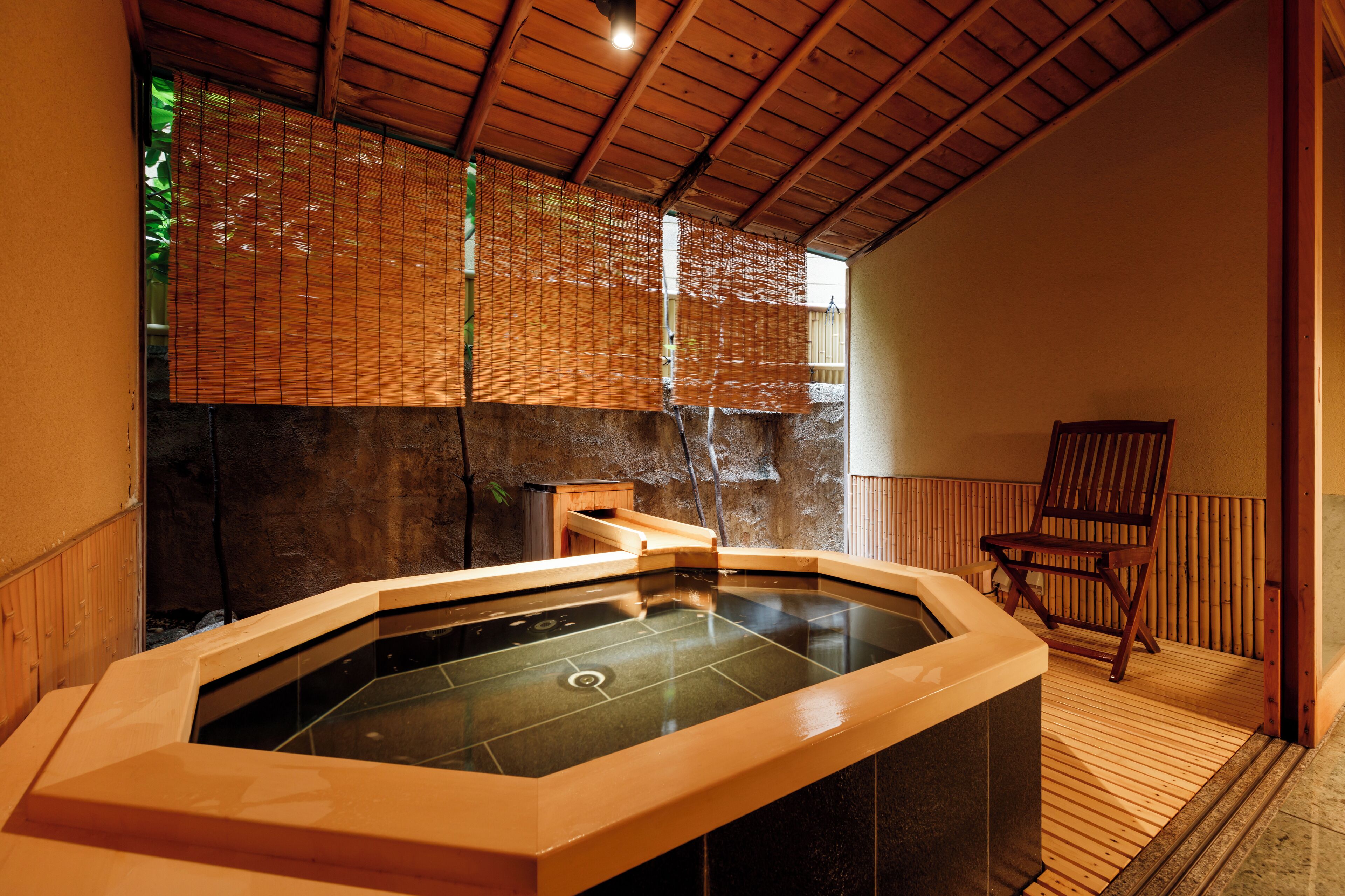 A hot tub with a wooden ceiling and walls, a wooden chair, and a stone wall in the background.