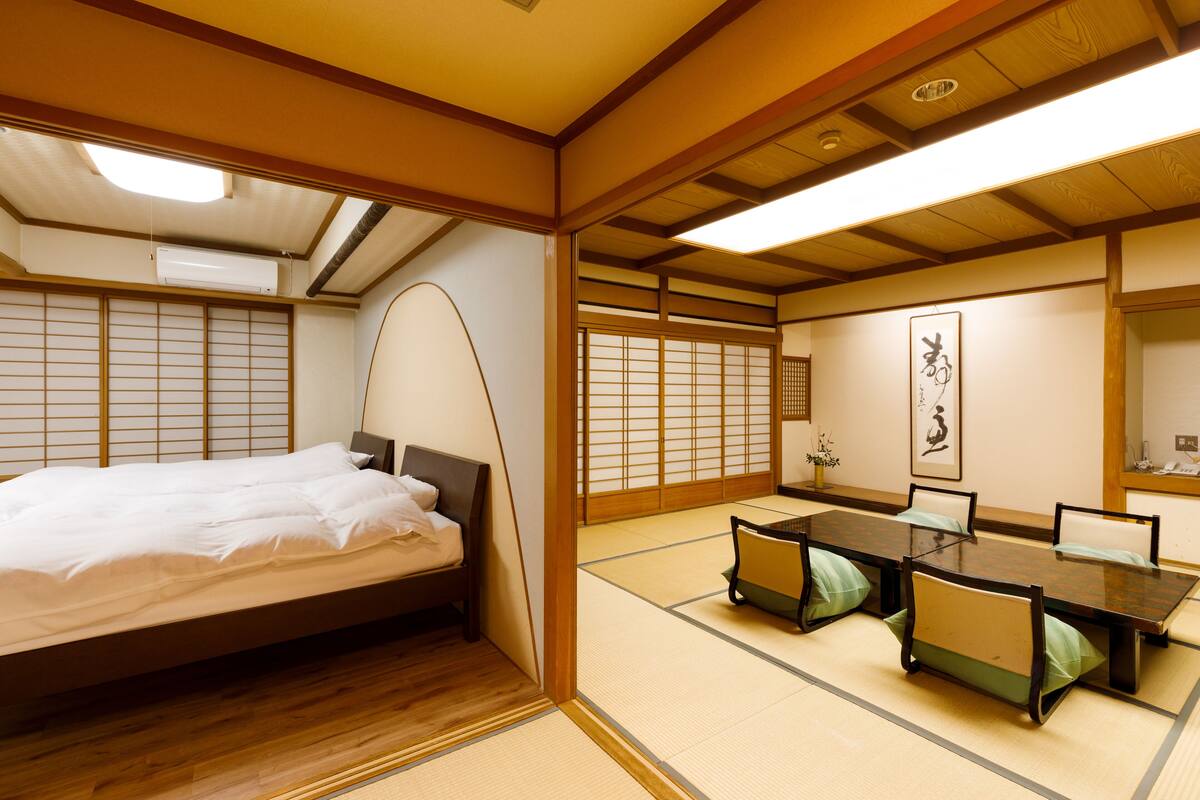 Una habitación japonesa tradicional con suelo de tatami, una cama baja de madera, una mesa de comedor con sillas y puertas correderas shoji.