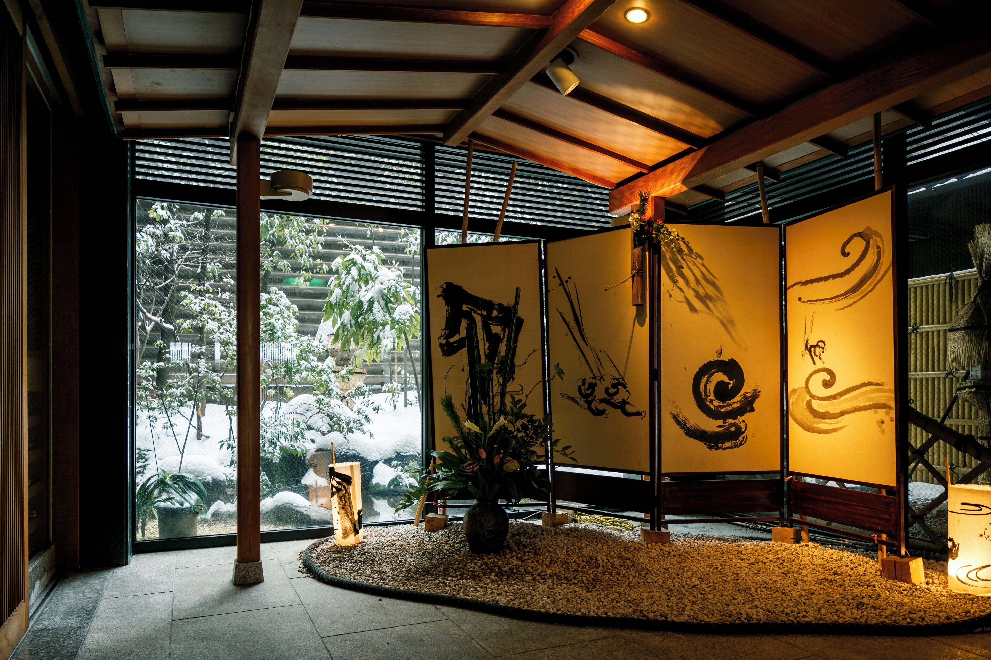 A traditional Japanese room with tatami flooring, sliding doors with calligraphy, and a view of a snowy garden.