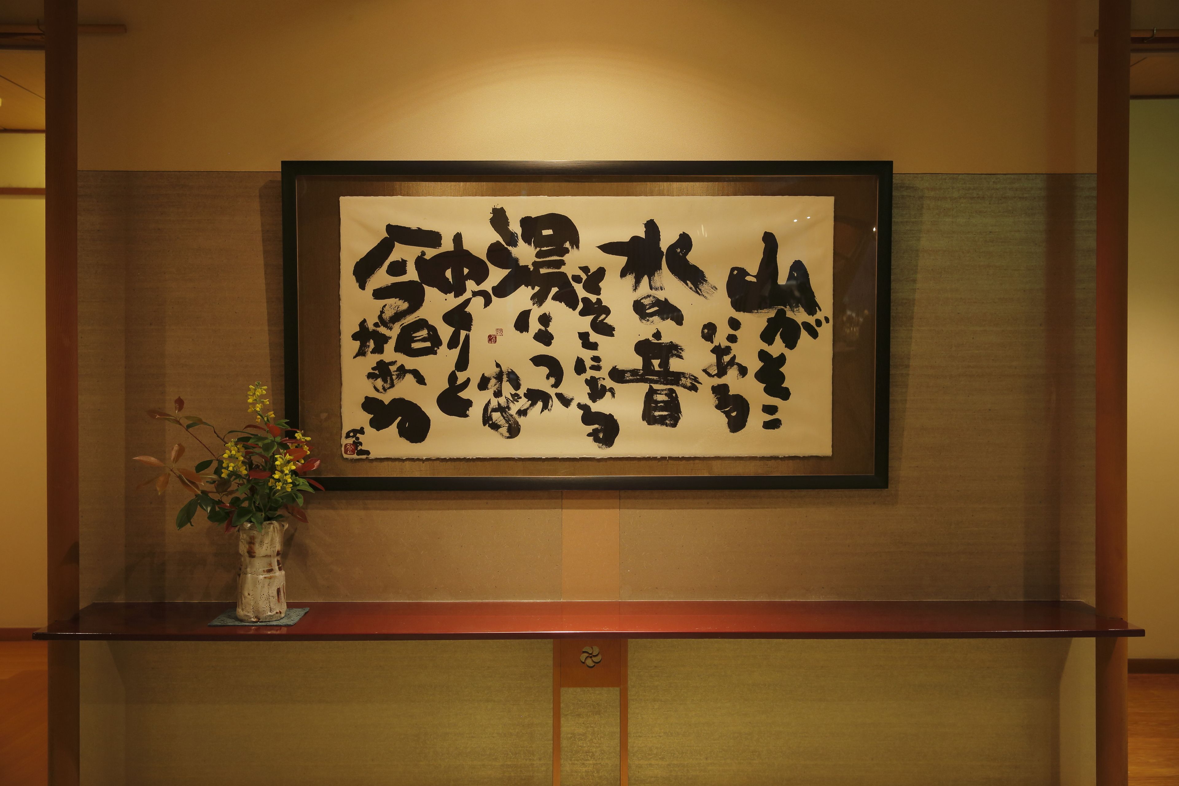 Ein gerahmtes Kalligraphie-Kunstwerk an der Wand, eine kleine Vase mit Blumen und ein Holzregal darunter.