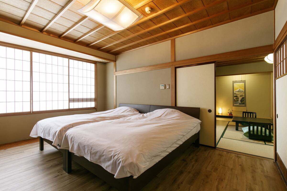 Una habitación con cama, suelo de madera, techo con vigas de madera y una puerta corredera que da a un comedor.