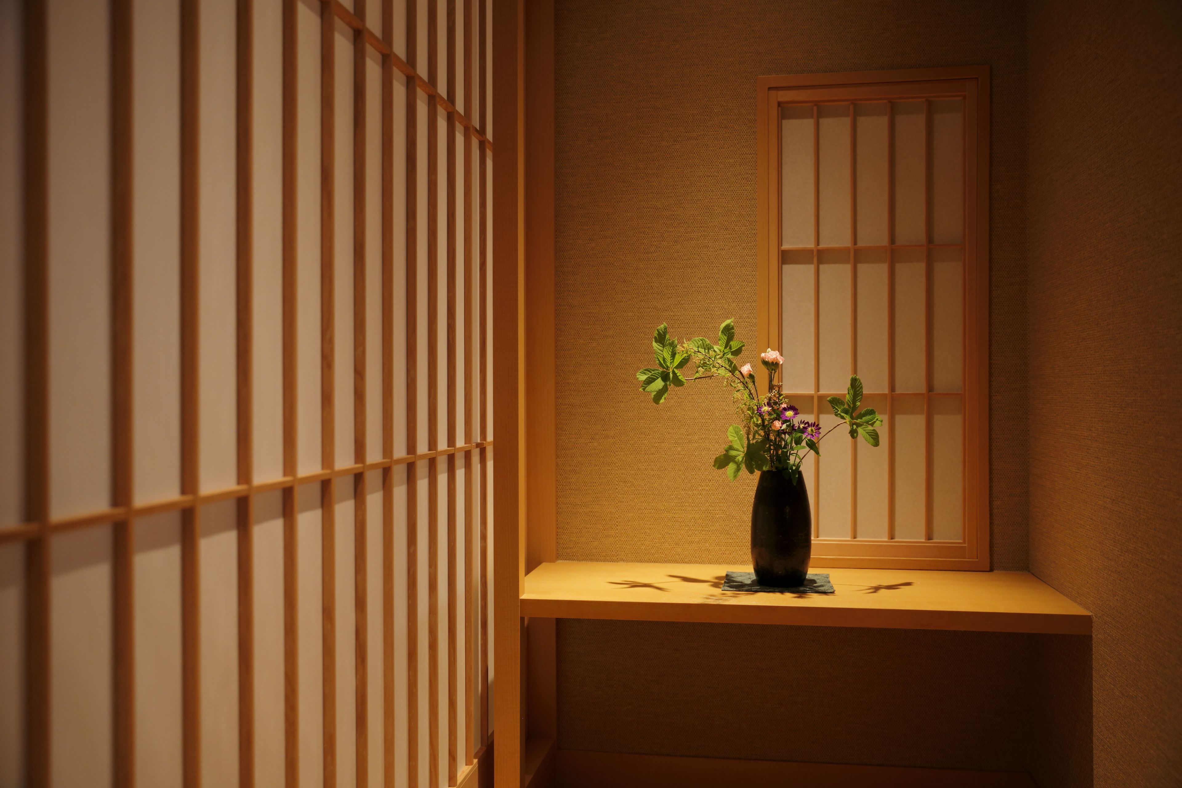 Una habitación japonesa tradicional con suelo de tatami, un shoji y una pequeña mesa con un jarrón de flores.