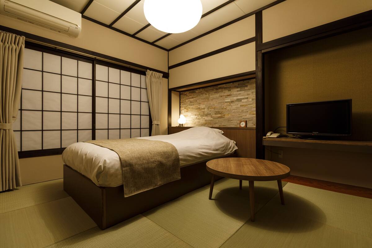 Una habitación con cama, una mesita, un televisor y un ventanal con cortinas.
