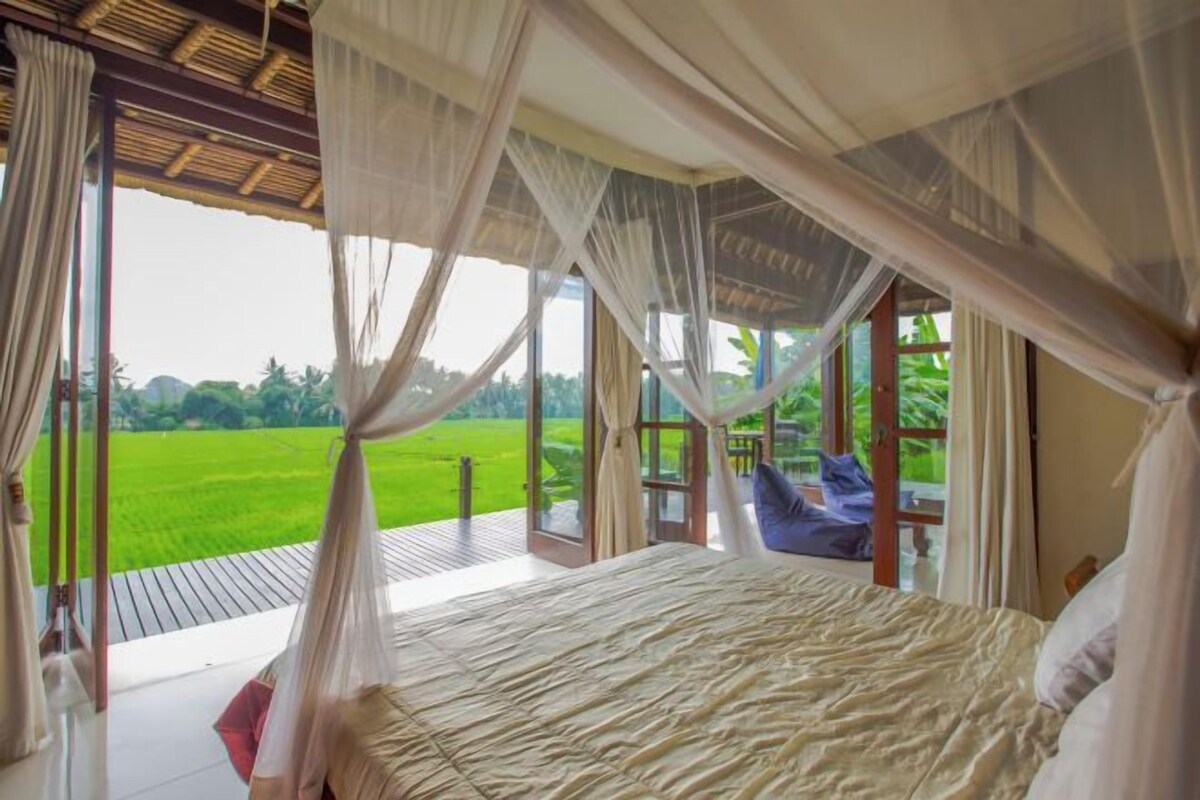 A bedroom with a large bed, a view of a green field, and a wooden deck outside.