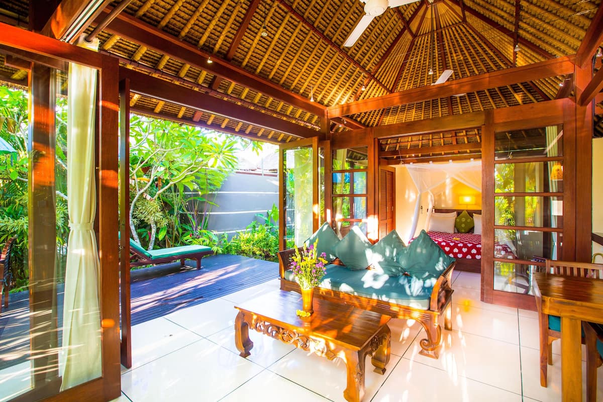 A spacious indoor area with a wooden ceiling, a sofa with cushions, a coffee table, and a glass door leading to an outdoor area with a bench and greenery.
