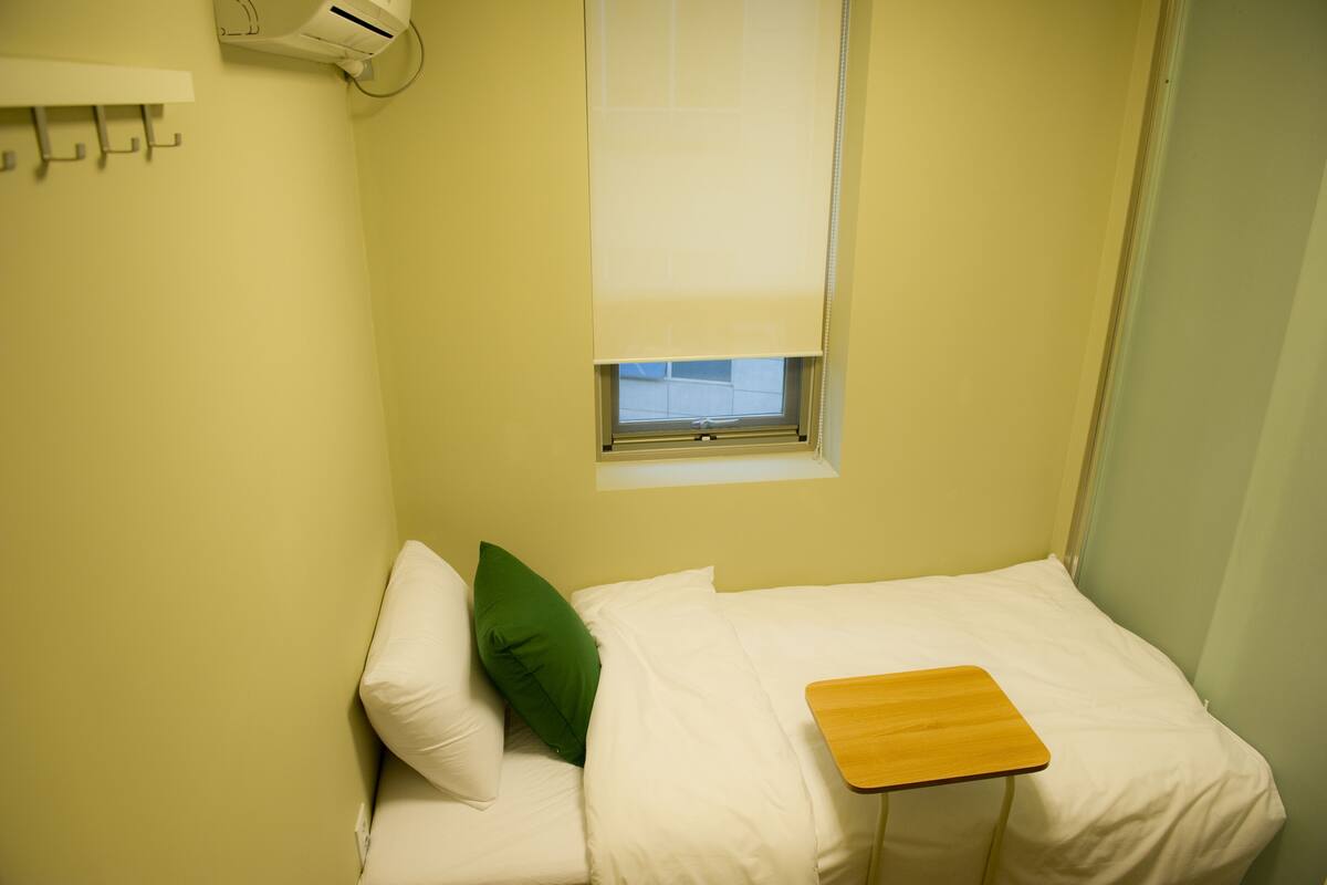 A small, single-bed room with a window, a bedside table, and a wall-mounted air conditioning unit.
