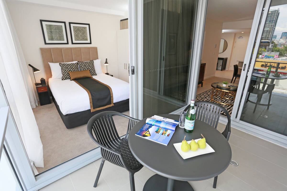 Una habitación de hotel con una cama, una mesa de comedor con sillas, una revista y dos peras.