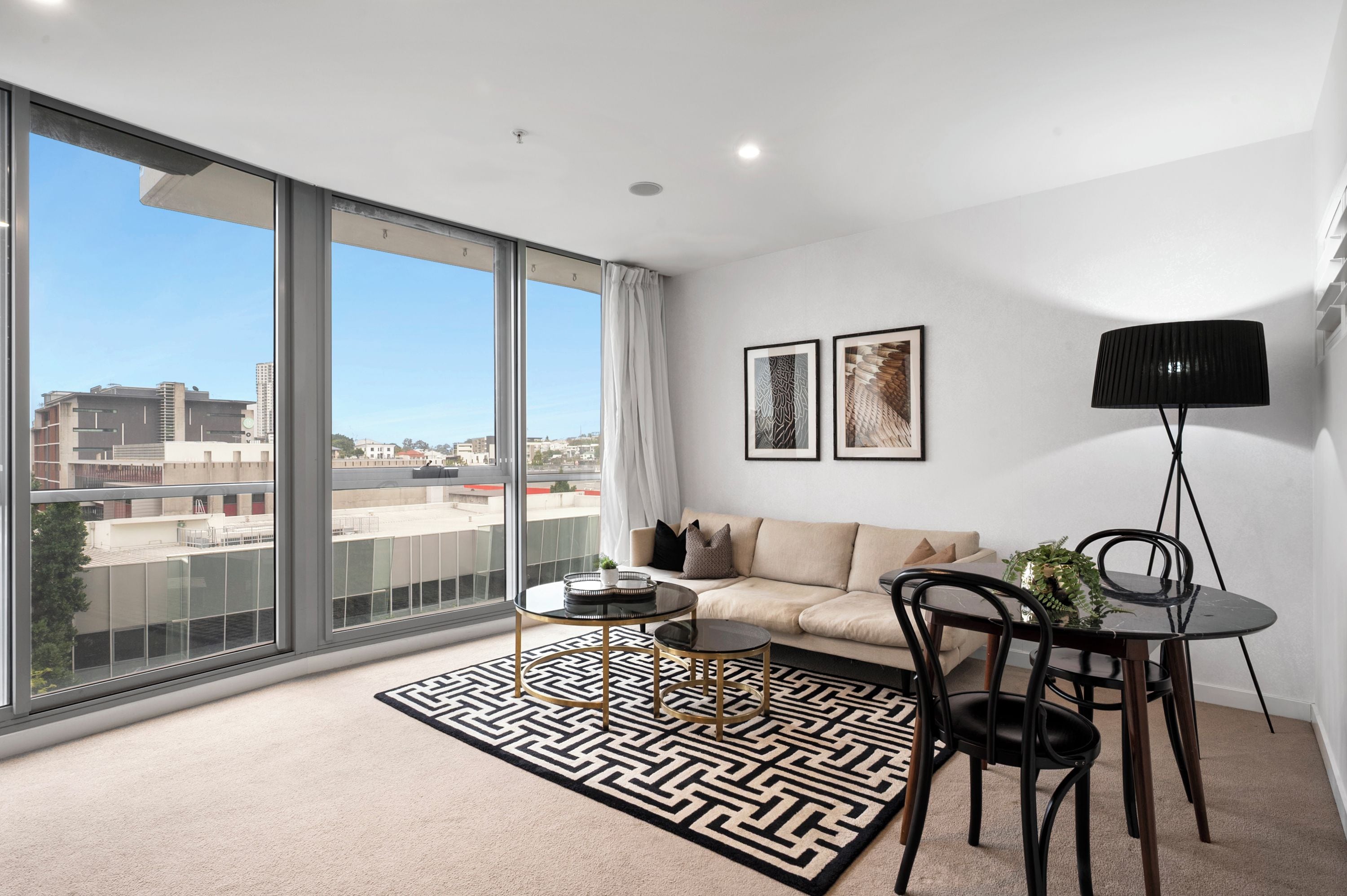 A modern living room with a large window, a sofa, a coffee table, a floor lamp, and two framed artworks.