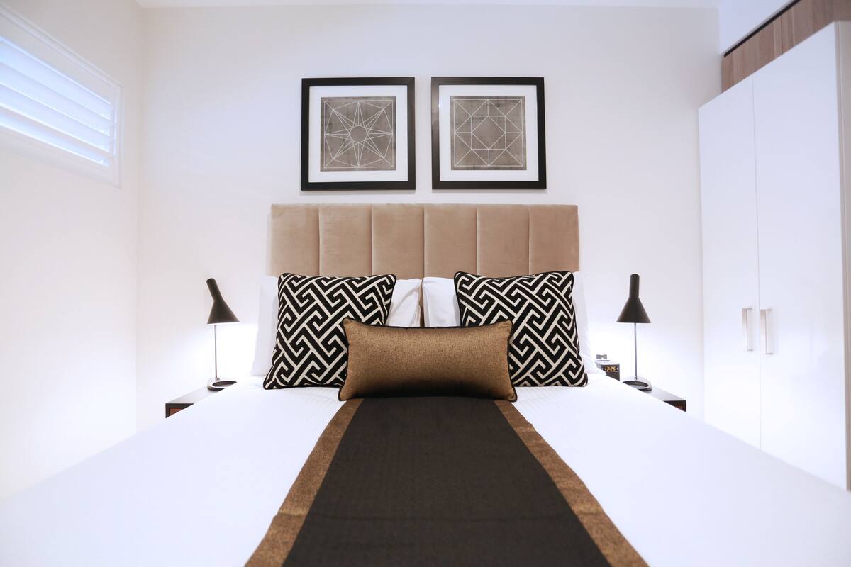 Un cuarto de hotel ordenado con una cama, dos cuadros geométricos en la pared, un cabecero y lámparas de noche.