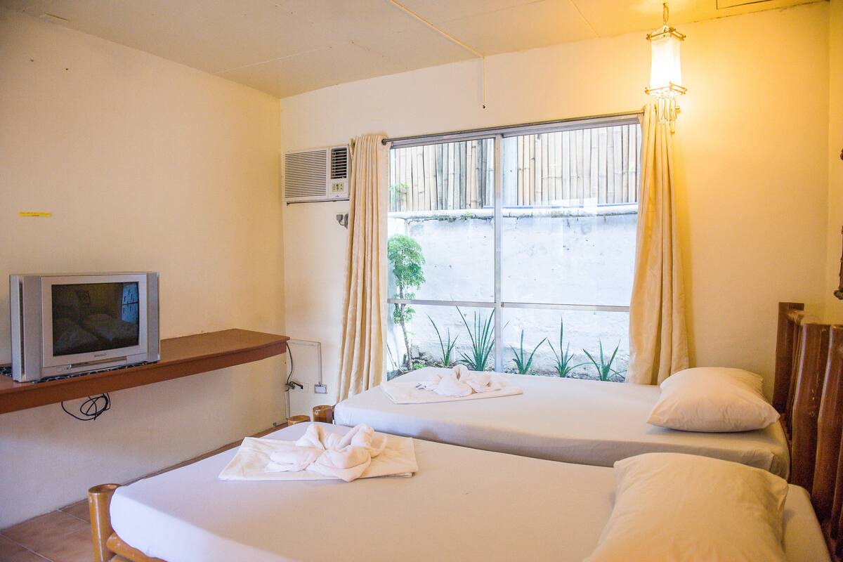 Une chambre d’hôtel avec deux lits, un téléviseur posé sur une étagère et une fenêtre avec des rideaux.