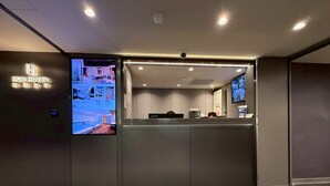 Zona de recepción de hotel con pantalla digital, mostrador de recepción y cámara de seguridad.